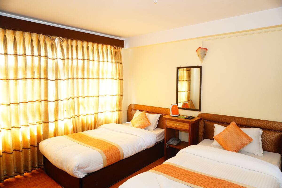 Quarto de hotel com duas camas, cabeceira de madeira, mesa de cabeceira com abajur e janela com cortinas.