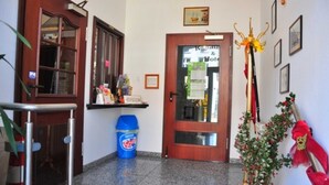 Un hall de hotel con una recepción, un bote de basura azul, una puerta con panel de vidrio y una planta decorativa con flores rojas.