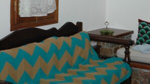Ein Zimmer mit einem raumhohen Fenster mit Holzrahmen, einem gemusterten Kissen auf einer Couch, einem kleinen Tisch mit einer grünen Schüssel und einem dekorativen Teller an der Wand.