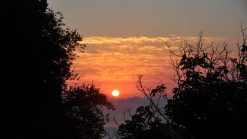 Een zonsondergang, waarbij de zon gedeeltelijk verborgen is achter wolken, met silhouetbomen en een vergezicht op een wateroppervlak.