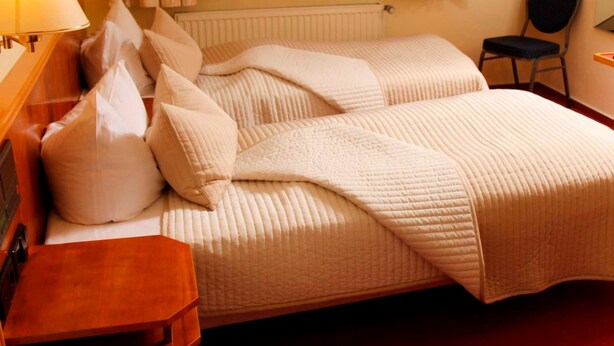 Habitación de hotel con dos camas, una mesita de noche de madera, una silla y un ventanal con cortinas.