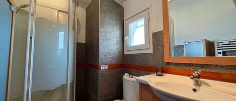 Een badkamer met een douchecabine van glas, een wit toilet, een wastafel met spiegel erboven en een raam.