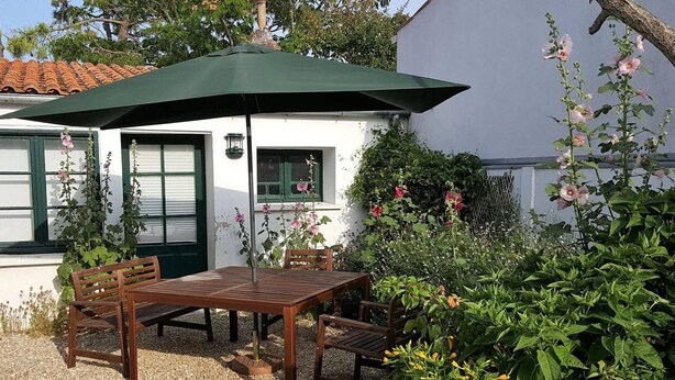 A wooden table and chairs under a green umbrella, set on gravel, with a white building and greenery in the background.