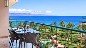 Un balcón con mesa lista para comer, con vista a una zona de piscina, palmeras y el océano.