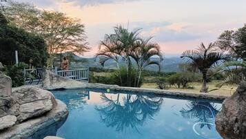 Una piscina con agua cristalina, rodeada de rocas y palmeras, con vistas a montañas distantes y un cielo nublado.