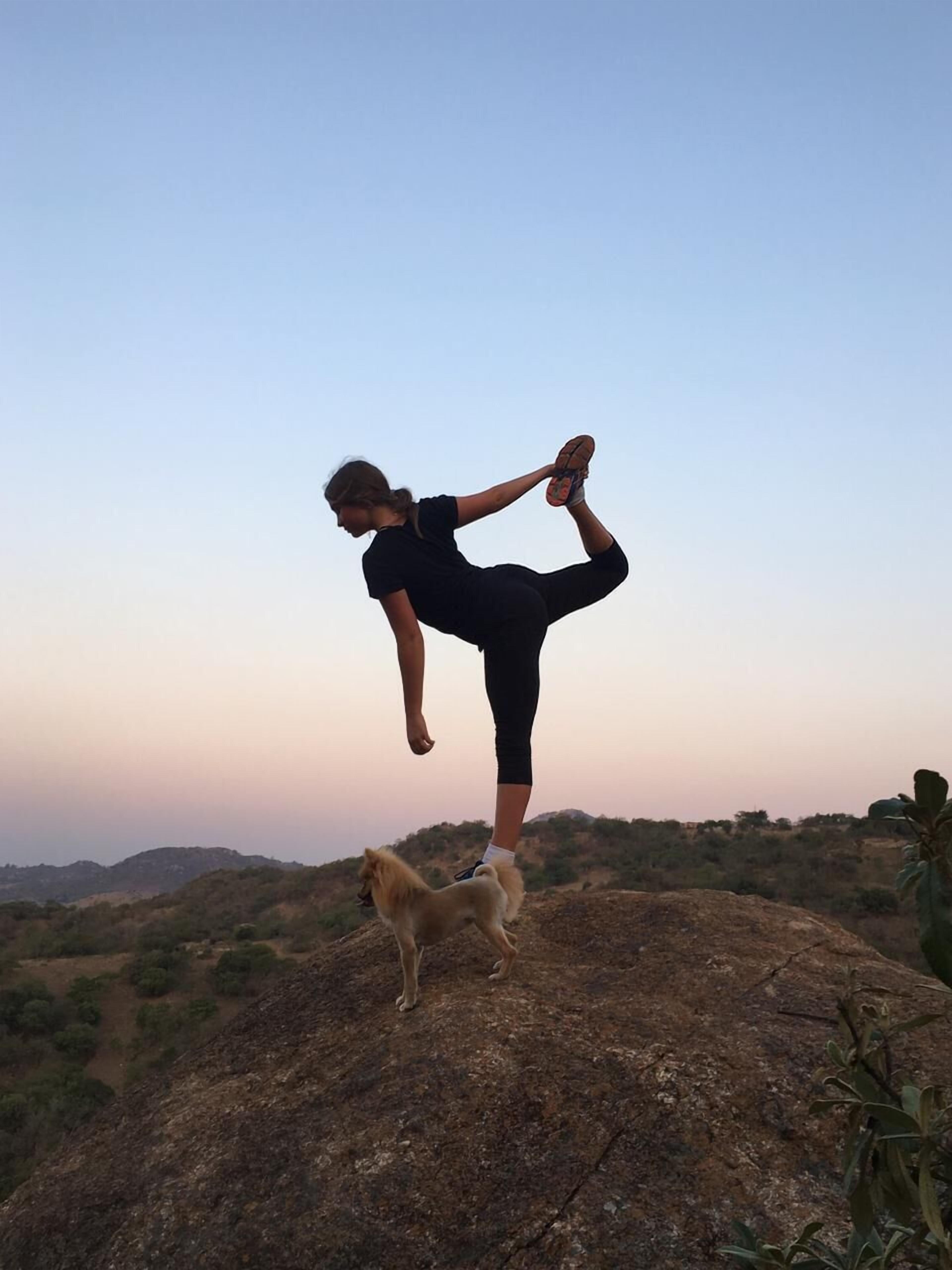 A person performing a yoga pose on a rocky outcrop with a dog nearby.