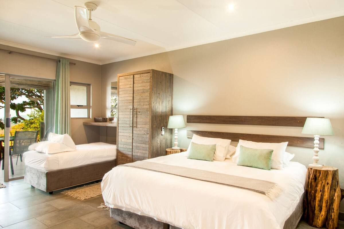 A bedroom with two beds, a ceiling fan, a wooden wardrobe, and a view of the outdoors.