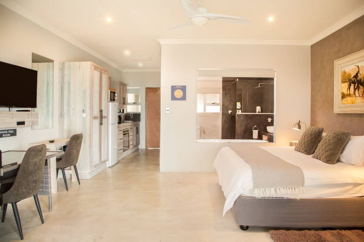 A modern hotel room with a bed, a desk with chairs, a kitchenette, and a bathroom with a large mirror.