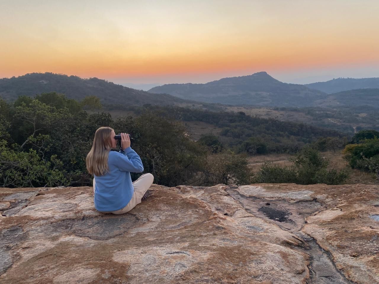 A person sitting on a rock, taking a photo of a landscape with hills and trees during sunset.