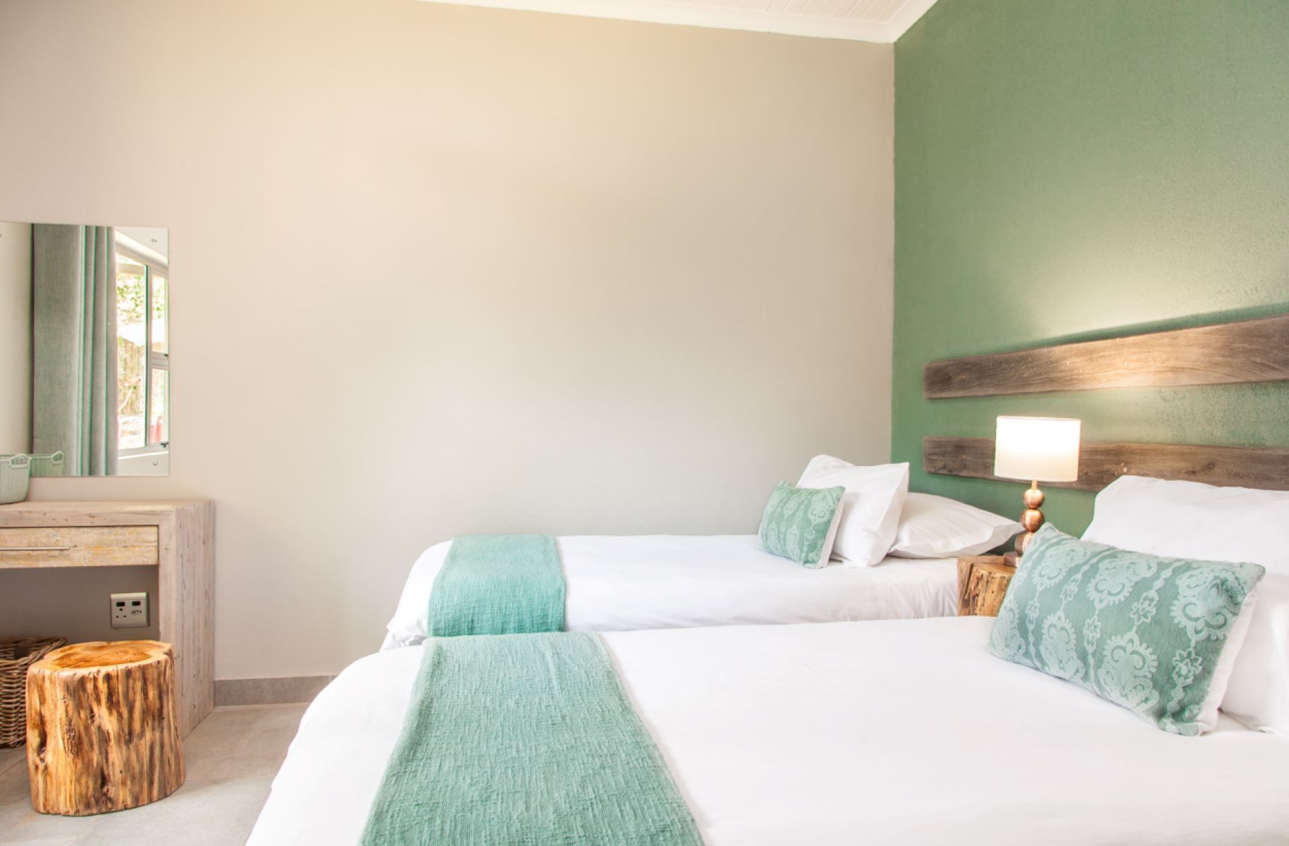 A hotel room with two beds, a wooden nightstand with a lamp, a small wooden stool, and a desk with a mirror.