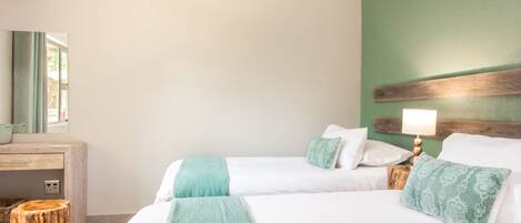 A hotel room with two beds, a wooden nightstand with a lamp, a small wooden stool, and a desk with a mirror.