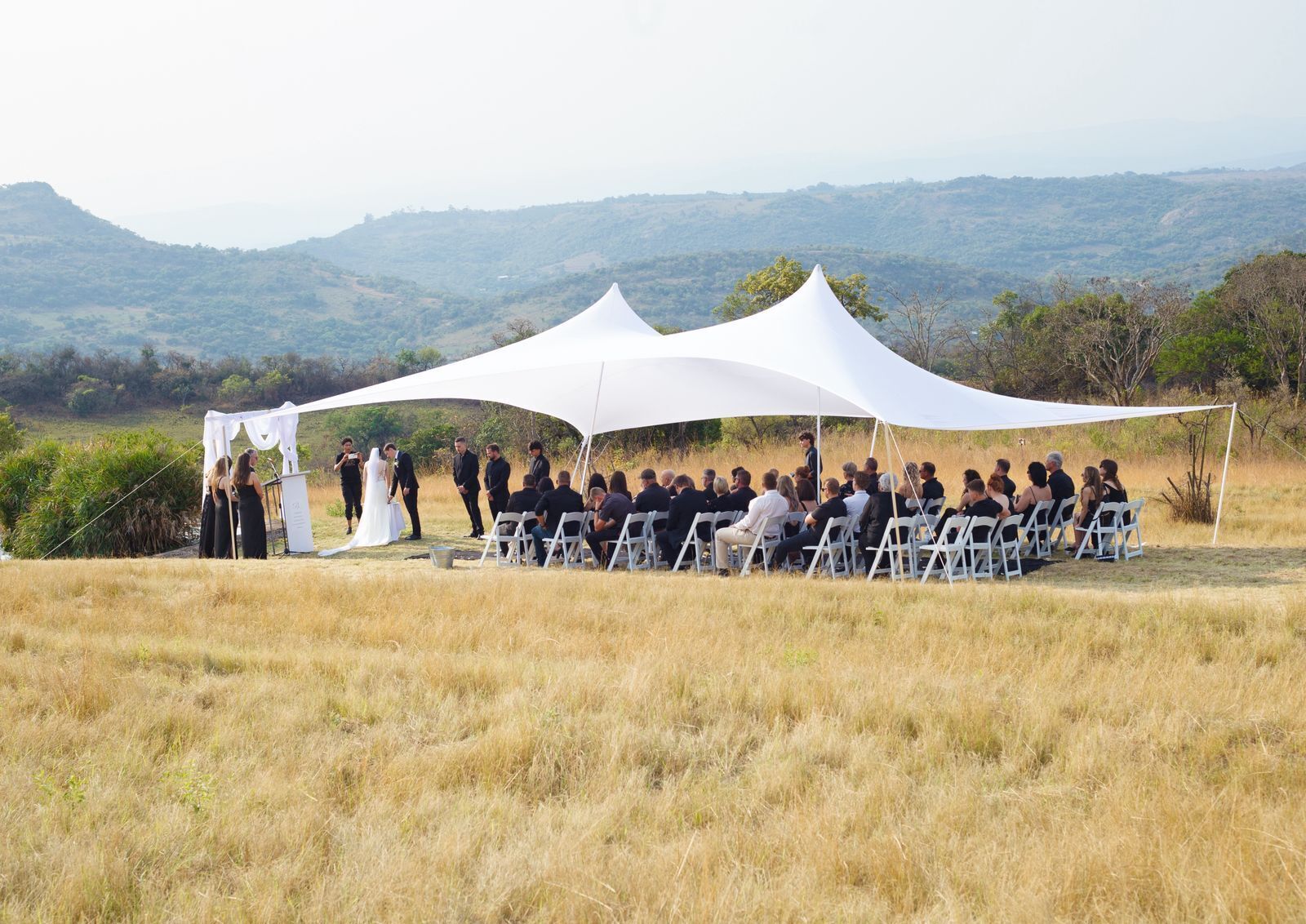An outdoor wedding ceremony with a white tent and guests seated on white chairs.
