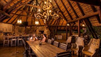 A rustic dining area with a wooden table and chairs, a stone wall, and a thatched roof.