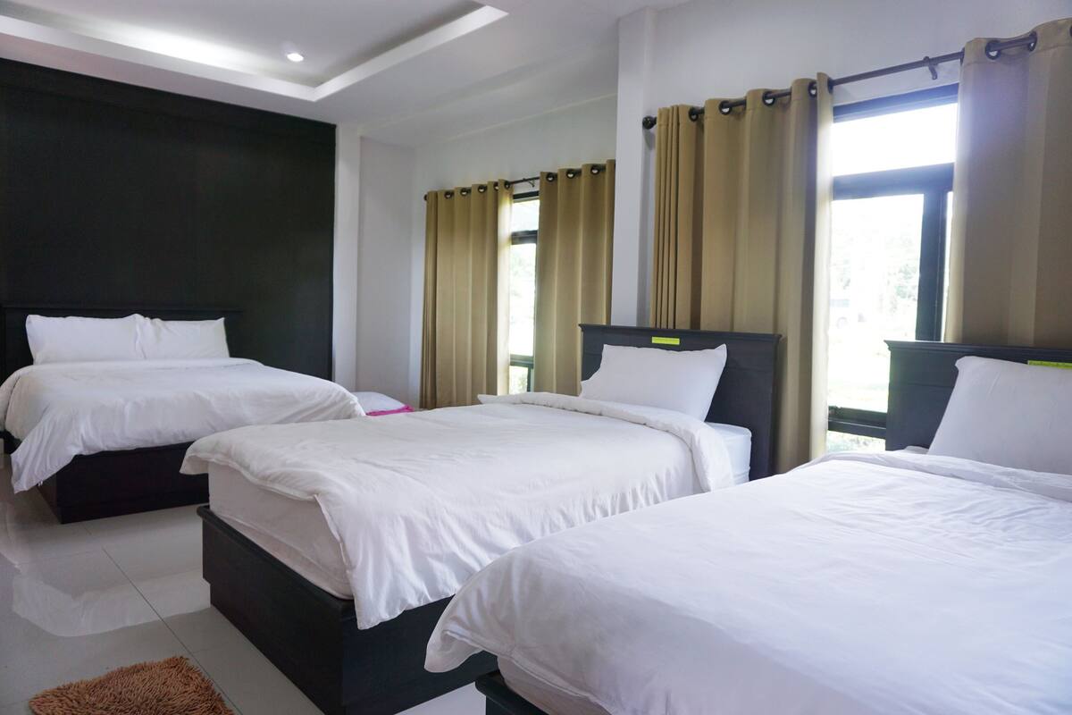 Ein Hotelzimmer mit drei Betten, einem dunkel gefärbten Kopfteil und einem großen Fenster mit Vorhängen.