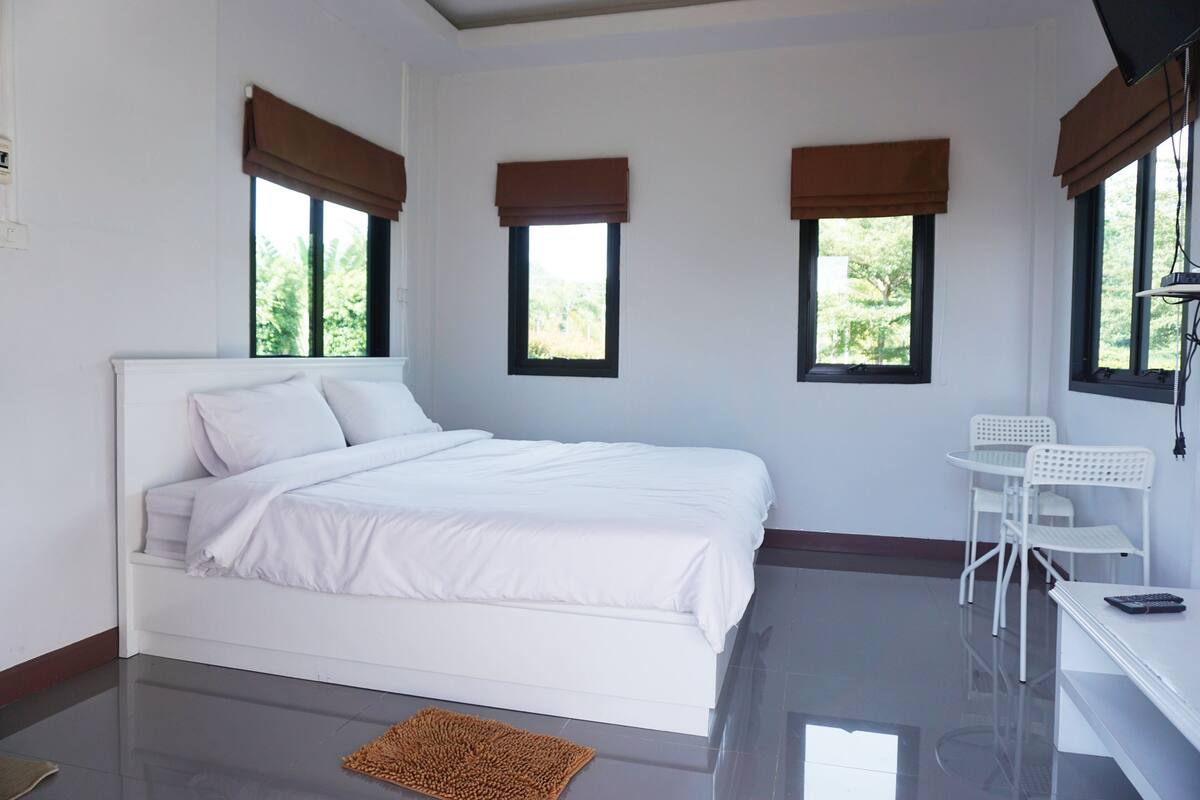 Ein Schlafzimmer mit einem weißen Bett, einem kleinen weißen Schreibtisch und drei Fenstern mit braunen Jalousien.