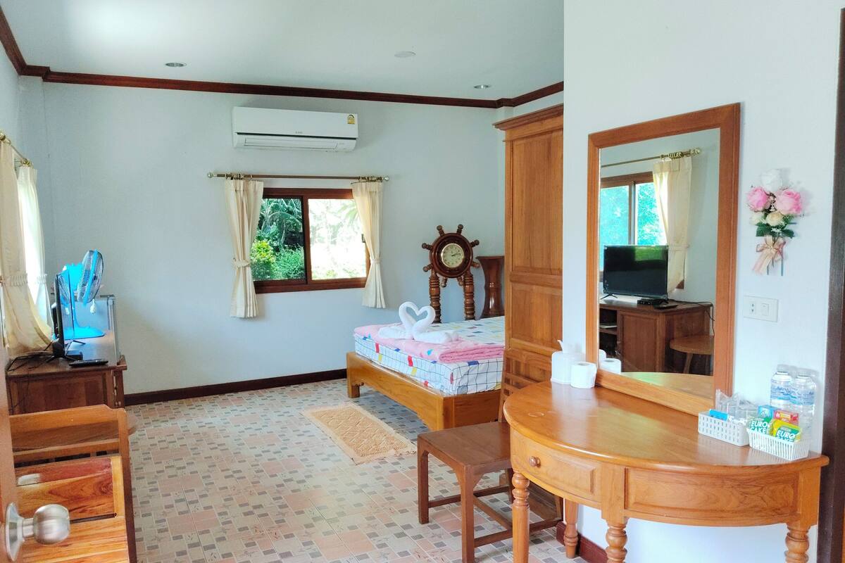 Un dormitorio con una cama, un aparador de madera, un escritorio, un espejo, un aire acondicionado, una ventana con cortinas, un reloj y un televisor.