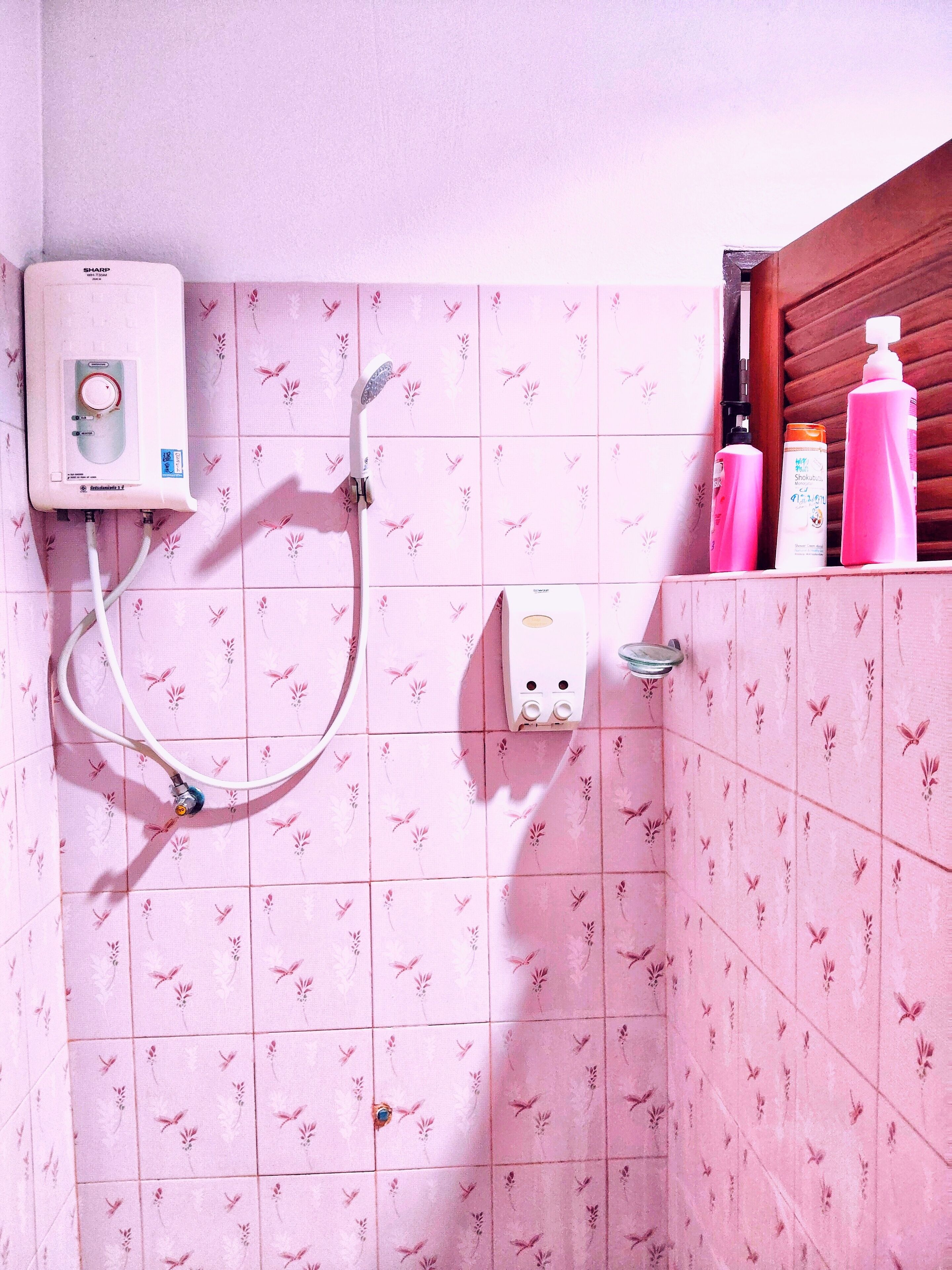 Une salle de bain avec des carreaux de céramique à motifs floraux roses, une douchette à main et des articles de toilette rangés sur une étagère.