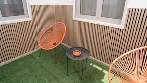 Un petit espace extérieur aménagé avec deux chaises orange et une table, entouré de lambris en bois verticaux et d’une fenêtre.