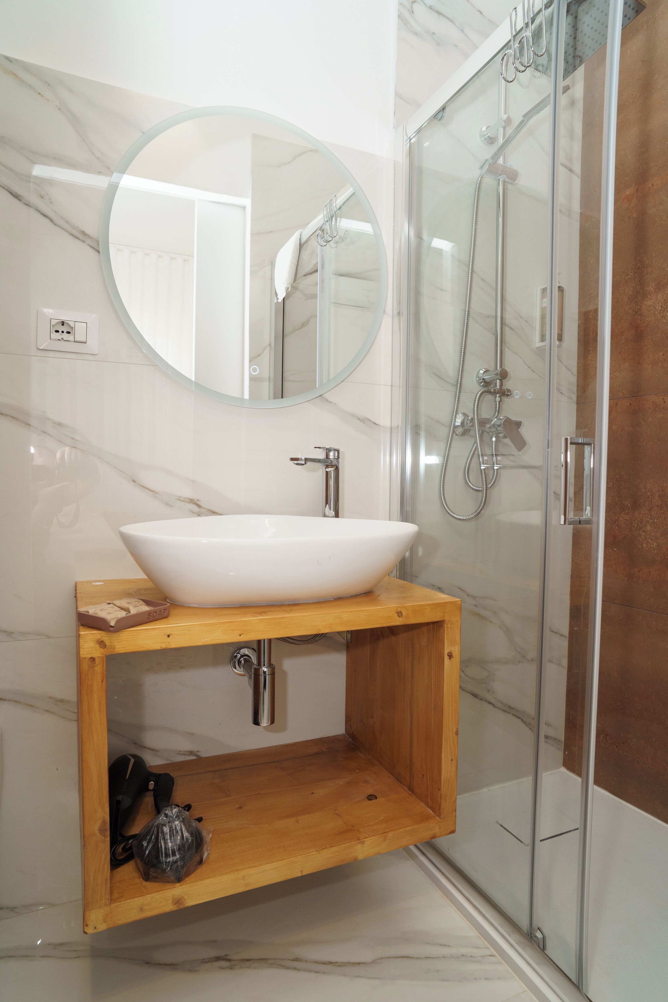 A bathroom with a white marble sink, a wooden vanity, a round mirror, and a glass-enclosed shower.