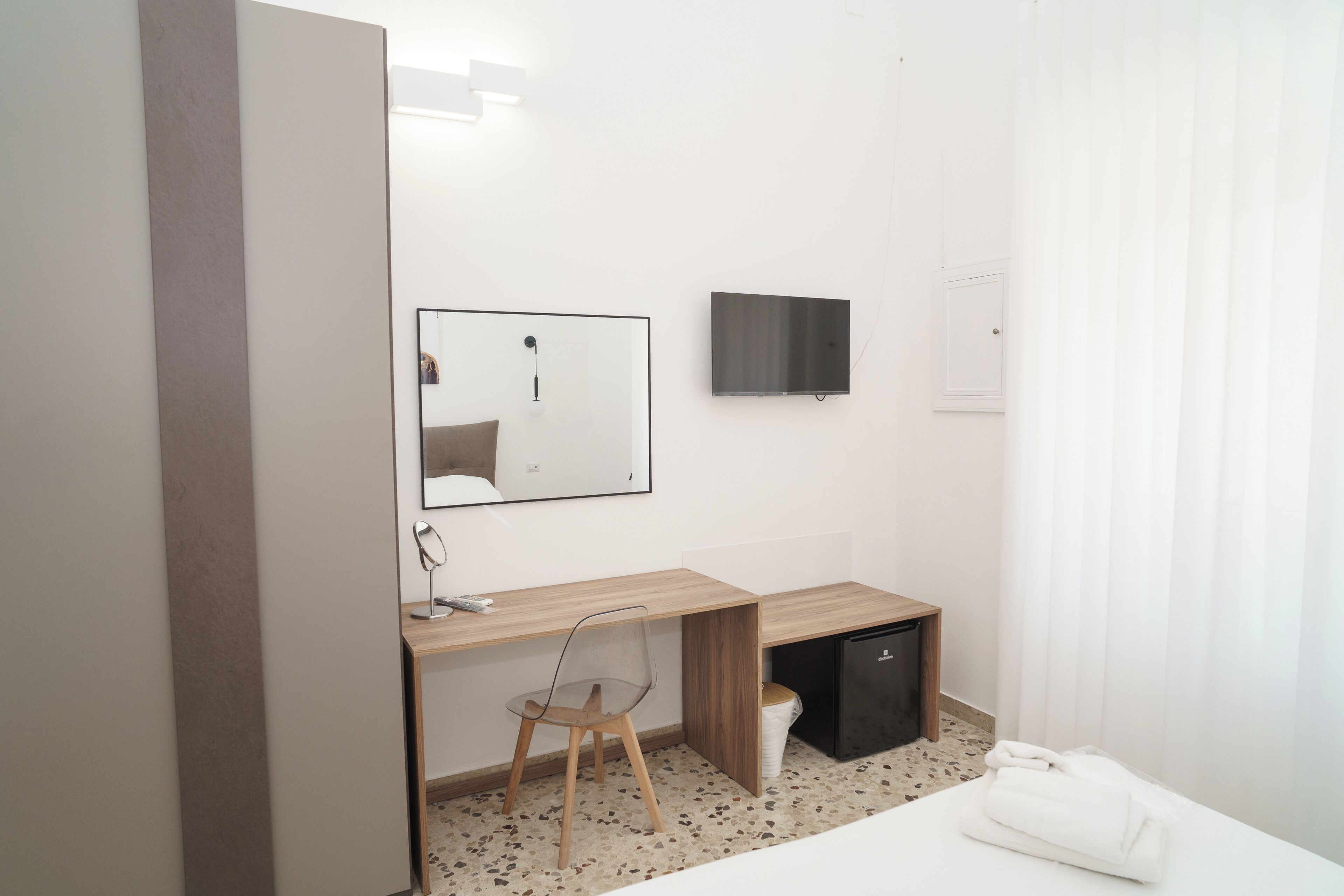 Une chambre d’hôtel comprenant un lit, un bureau avec une chaise, un petit réfrigérateur noir, un miroir et un téléviseur à écran plat fixé au mur.