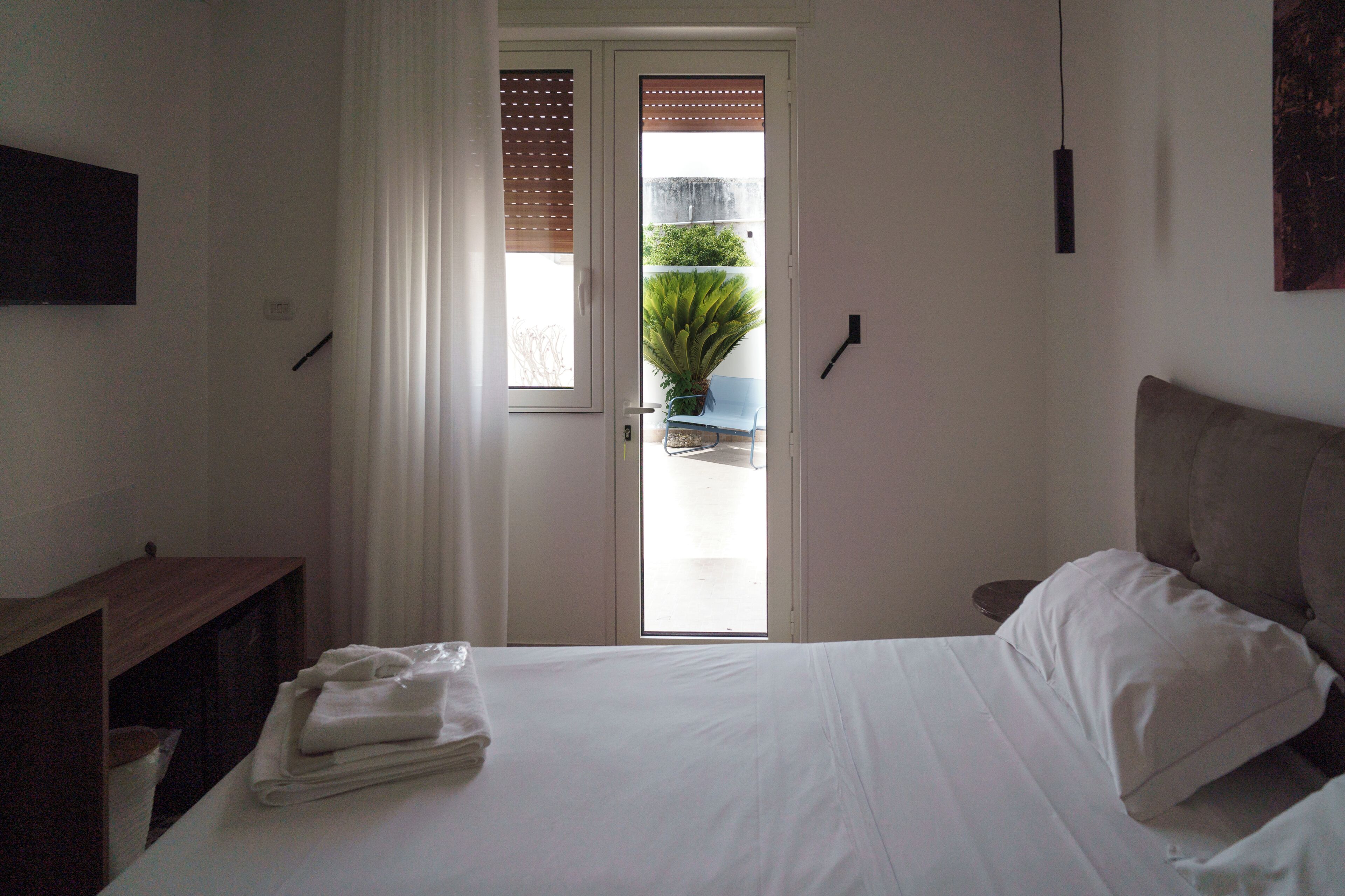 A bedroom with a bed, a nightstand, a window with blinds, and a view of the outdoors.