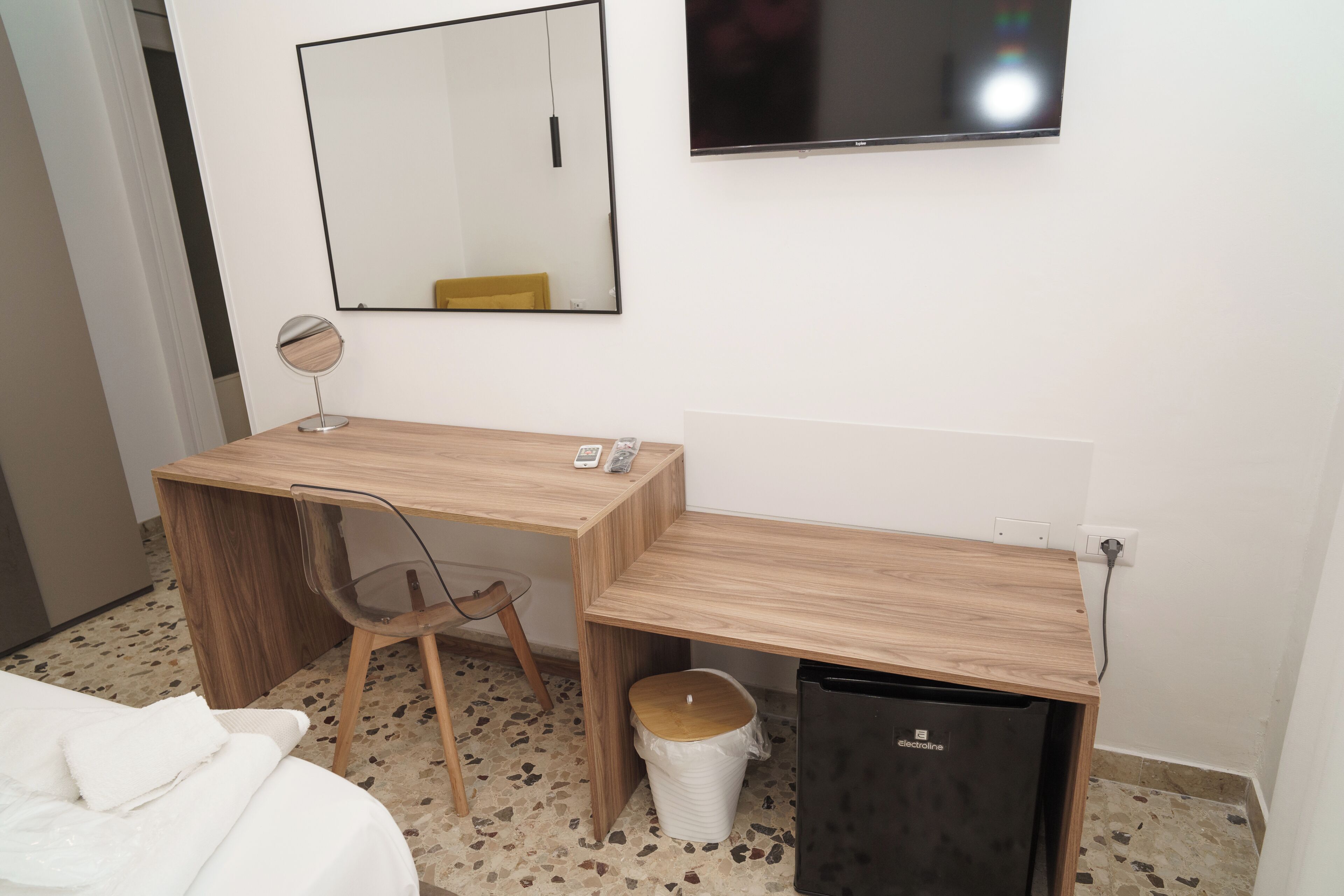 A hotel room with a wooden desk, a chair, a small trash bin, a mirror, and a television mounted on the wall.