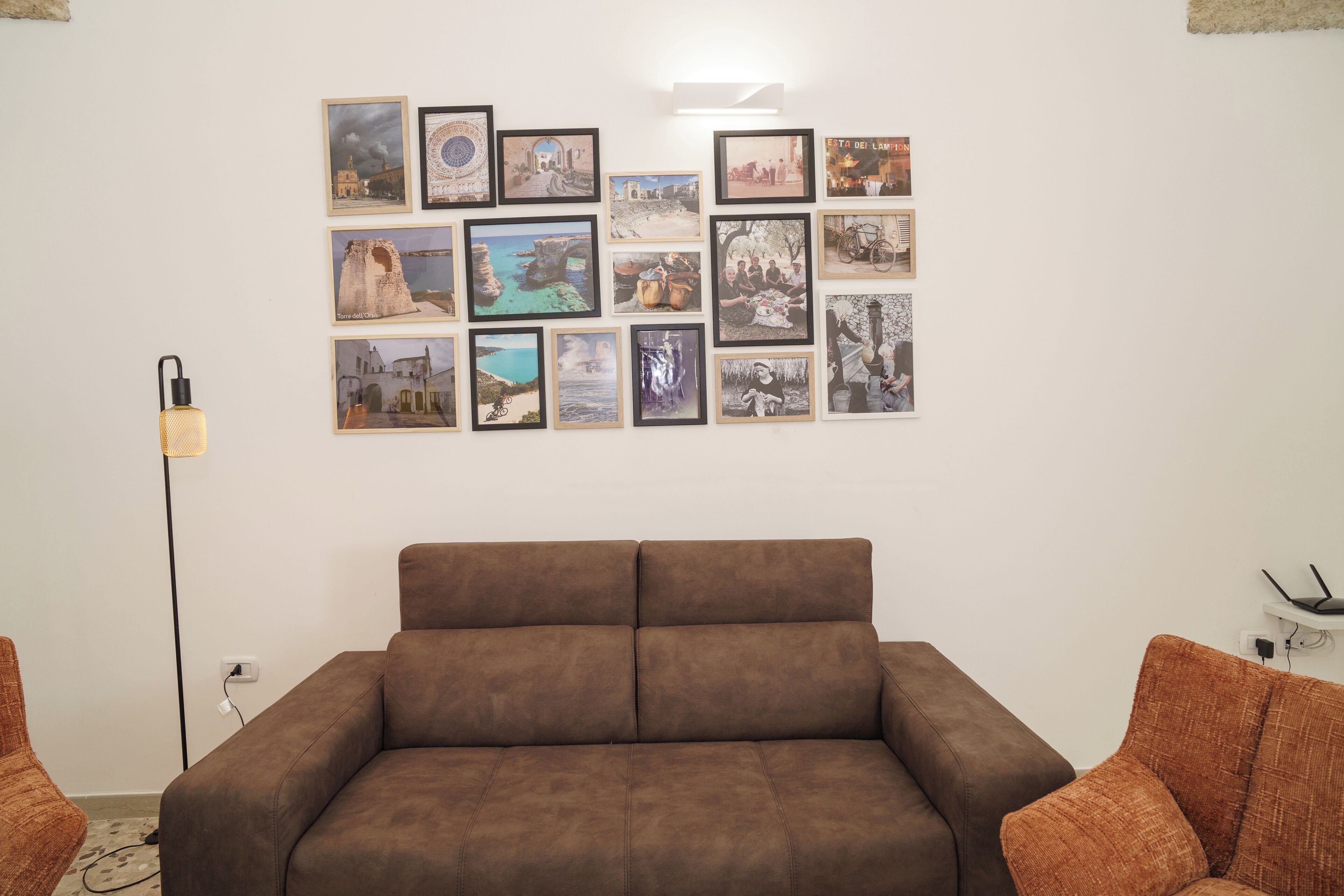 Una habitación con un sofá marrón, una lámpara de pie y una pared decorada con varias fotografías enmarcadas.