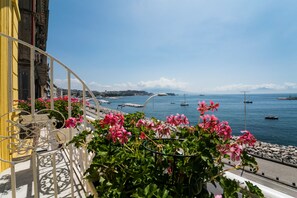 A balcony with pink flowers, a railing, and a view of the sea with boats.