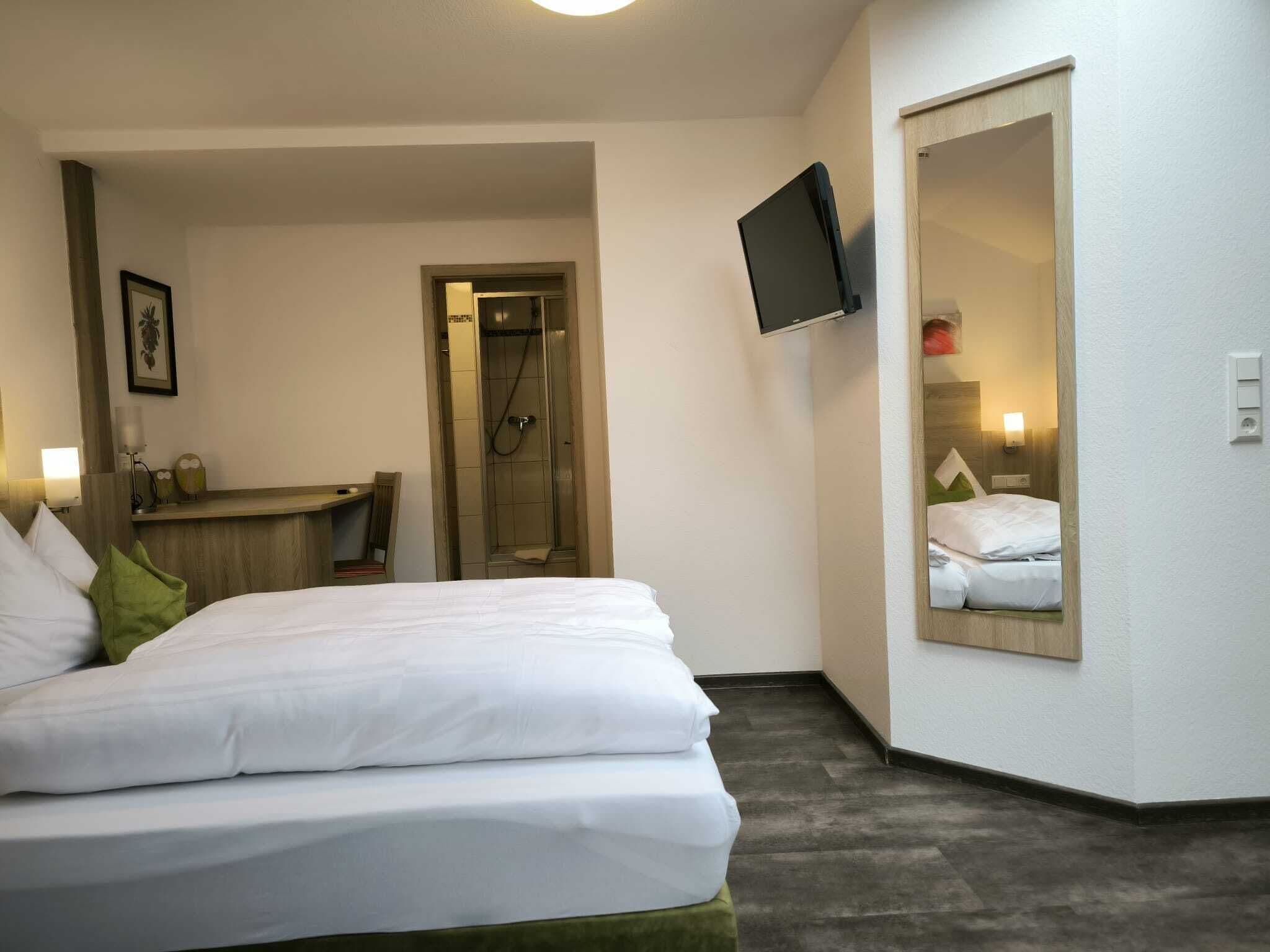 Ein Hotelzimmer mit Bett, Dusche, an der Wand montiertem Fernseher und Spiegel, der das Bett reflektiert.