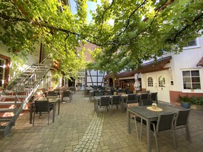 Ein Straßencafé mit Tischen und Stühlen auf Kopfsteinpflaster, umgeben von Bäumen und Gebäuden.