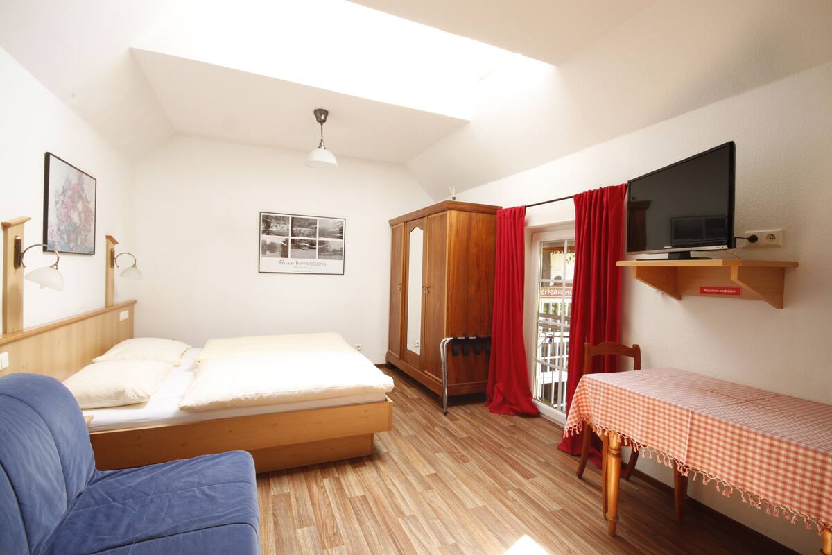 A room with a bed, a chair, a table with a checkered tablecloth, a TV on a shelf, a wardrobe, and a ceiling light.