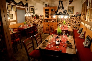 A cozy dining area with a wooden table set for four, surrounded by red upholstered chairs. A wooden cabinet with glass doors is visible in the background, and a stone wall with stacked firewood adds to the rustic ambiance. - Am Treidelpfad (Hassmersheim)