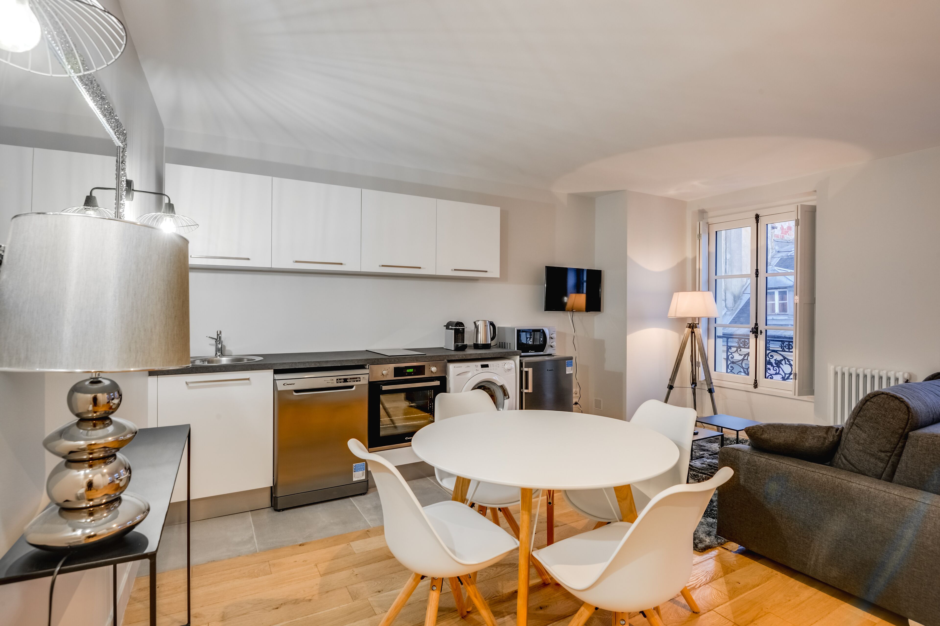 A compact kitchen with modern appliances, a dining area with white chairs, and a sofa.