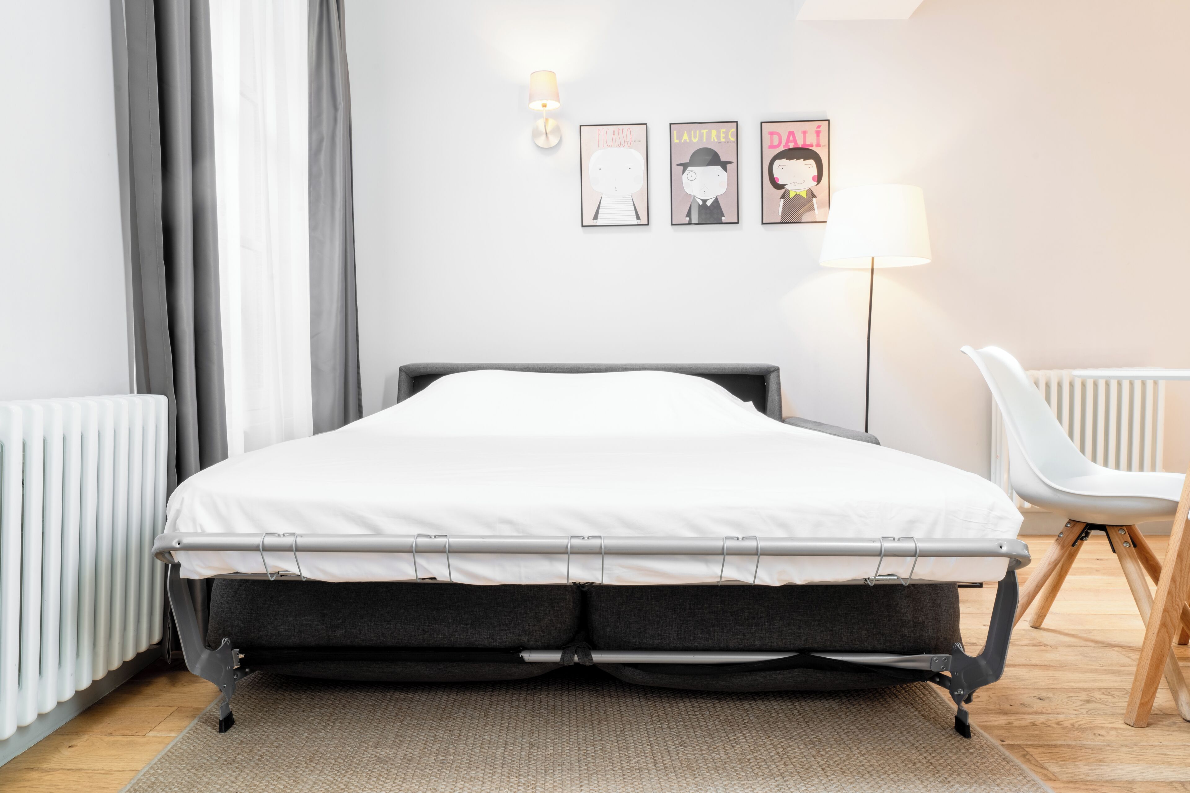 Een net opgemaakt bed met een grijze matras en witte lakens, een witte radiator, een grijze gordijn en een witte stoel.