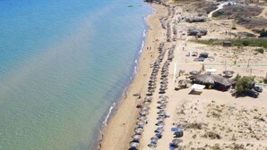 An aerial view of a beach with rows of umbrellas and beach chairs, a sandy shore, and buildings in the background. - Villa Lobos (Zakynthos)