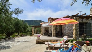 A stone building with a covered patio, two lounge chairs, and a colorful umbrella. - Villa Lobos (Zakynthos)