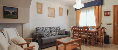 Ein Wohnzimmer mit einer Couch, Sesseln, einem Couchtisch und einem Esstisch mit Stühlen. Es gibt eine Chandelier, ein Bild an der Wand und ein Fenster mit Vorhängen.
