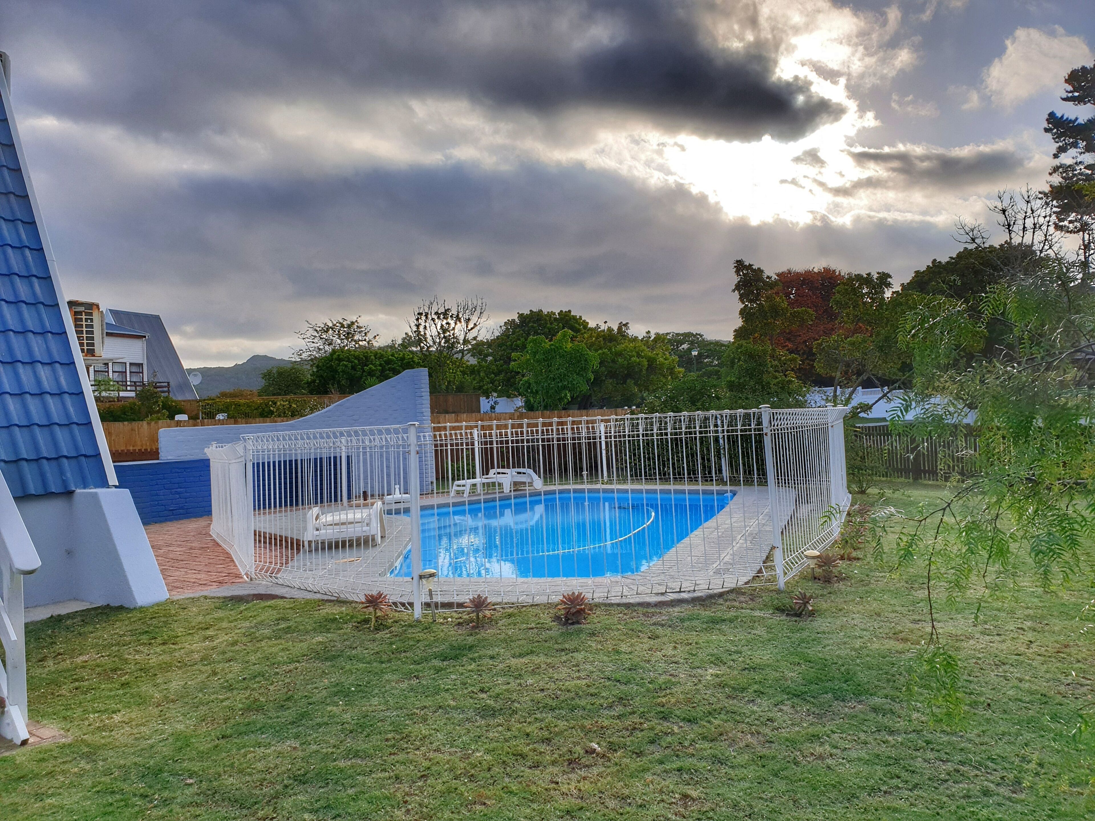 Una piscina con valla blanca, rodeada de césped y árboles, bajo un cielo nublado.