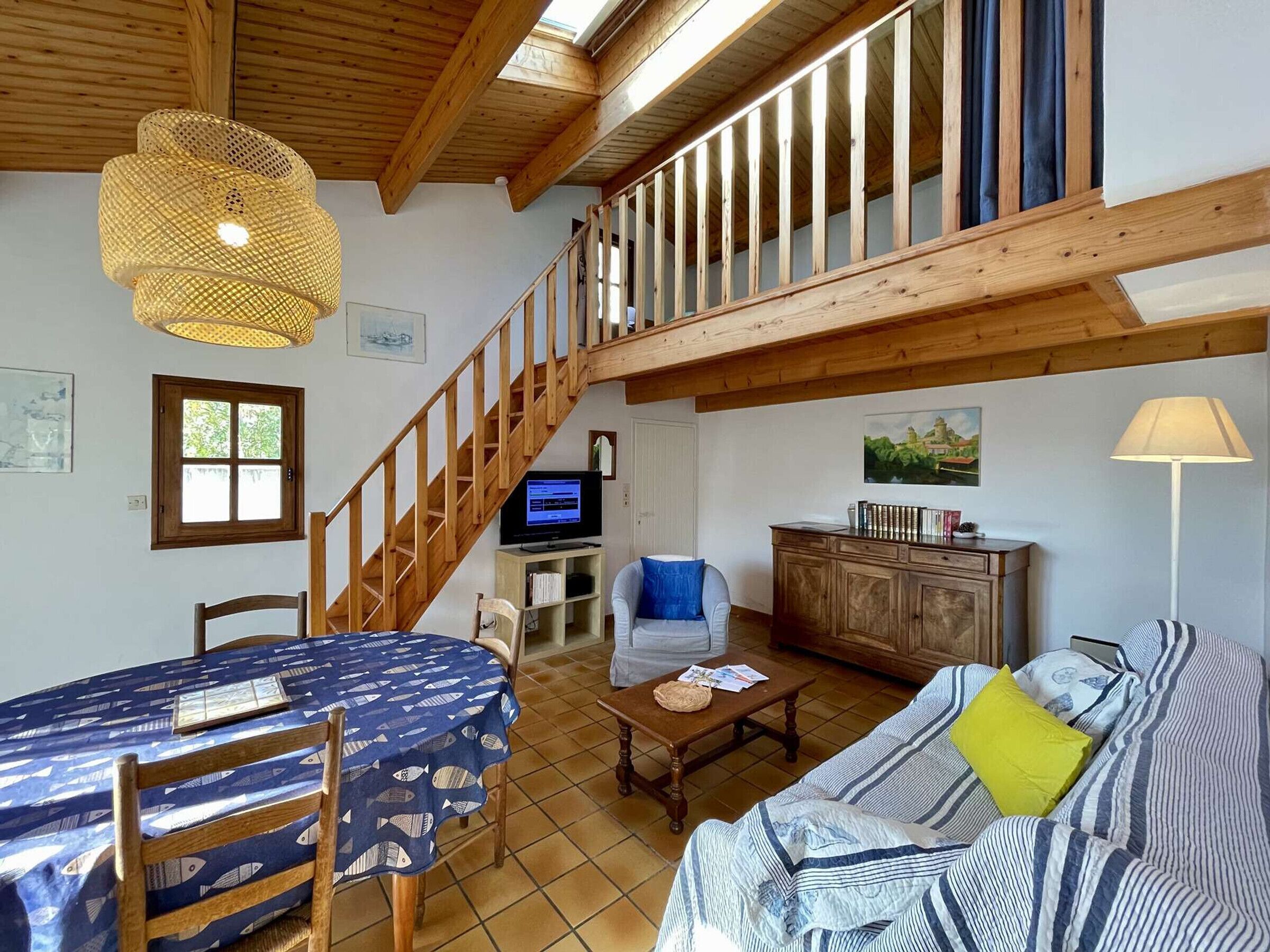 A spacious living area with a wooden staircase, a dining table with a blue tablecloth, a sofa with striped cushions, and a television in the background.