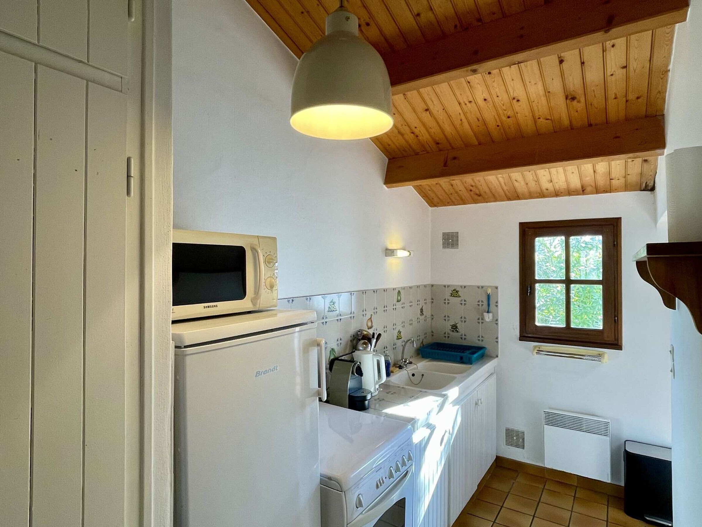 A compact kitchen with a microwave, Brandt refrigerator, sink, and a window with a view of greenery.