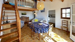 A rustic interior with a wooden staircase, a dining table with a fish-patterned tablecloth, and a kitchen area visible through an open door.