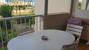 A balcony with a round table and white chairs, overlooking a garden and a beach.