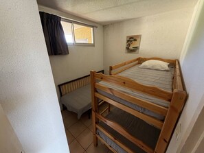 Une petite chambre avec un lit simple et un lit superposé en bois, une fenêtre avec des rideaux, un radiateur et un tableau accroché au mur.