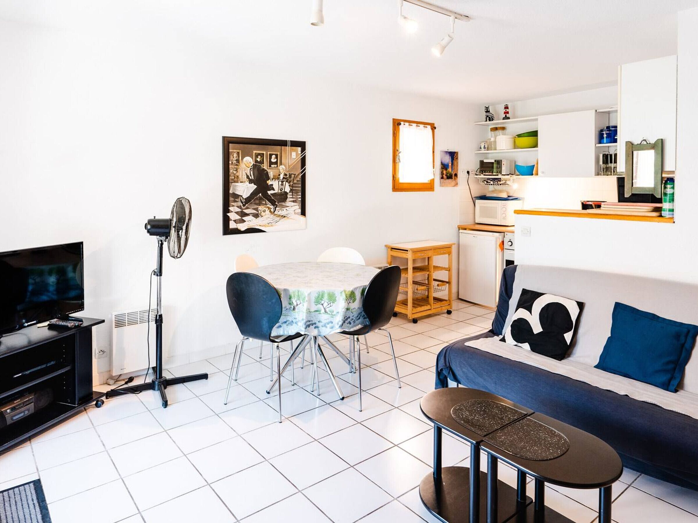 A compact living space with a TV, a fan, a dining table, a sofa, and a kitchenette.