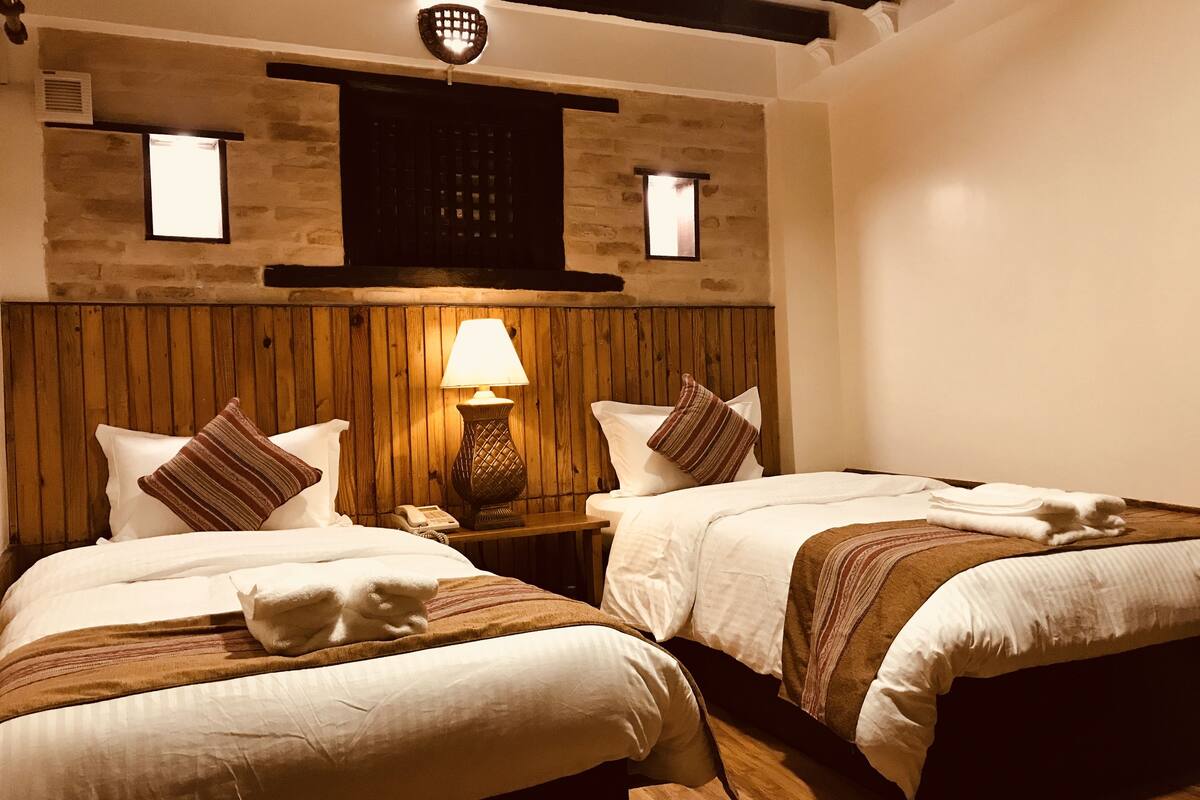 A hotel room with two beds, a wooden headboard, a nightstand with a lamp, and a small window.