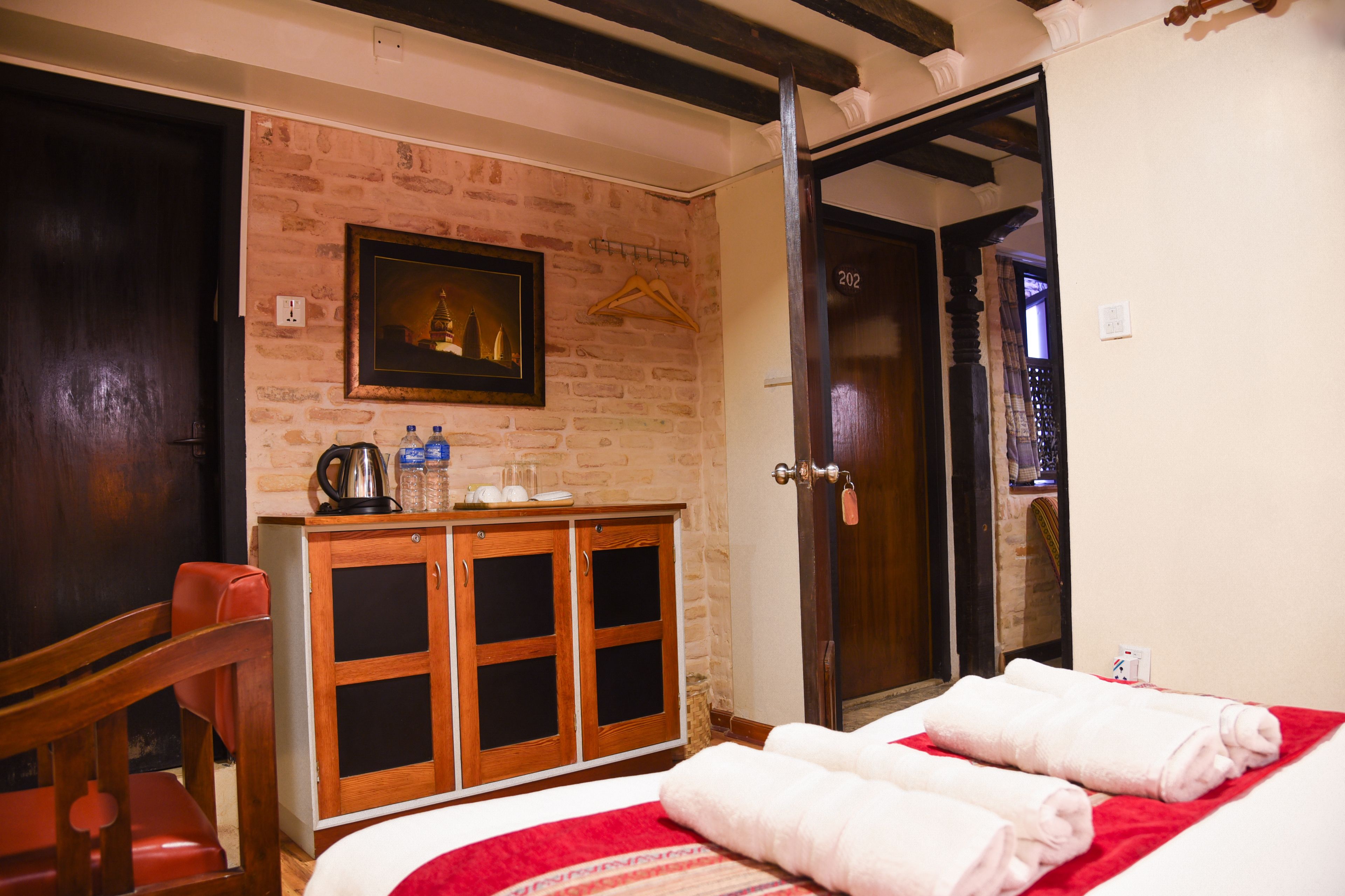 A hotel room with a brick wall, a wooden cabinet, a bed with red bedding, and a door leading to another room.
