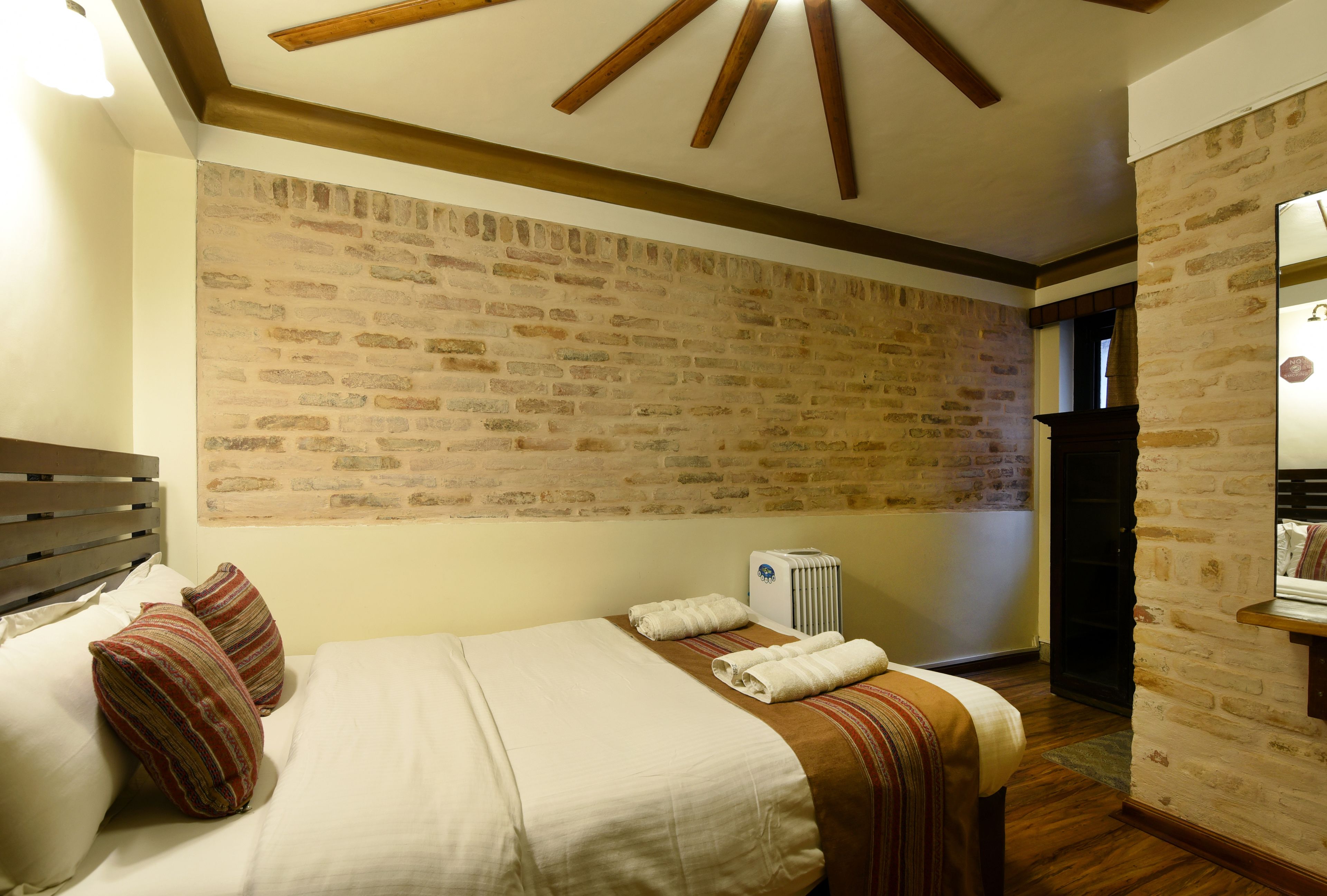Une chambre d’hôtel avec un mur en briques, des poutres en bois au plafond, un lit avec des oreillers, un radiateur et une petite table avec un miroir.