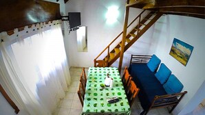 Un comedor con una mesa cubierta por un mantel verde, sillas de madera, un sofá con cojines azules, una escalera de madera y un televisor montado en la pared.