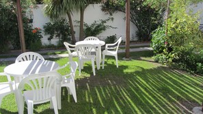 Un patio con una mesa y sillas blancas, rodeado de césped cuidado y diversas plantas.