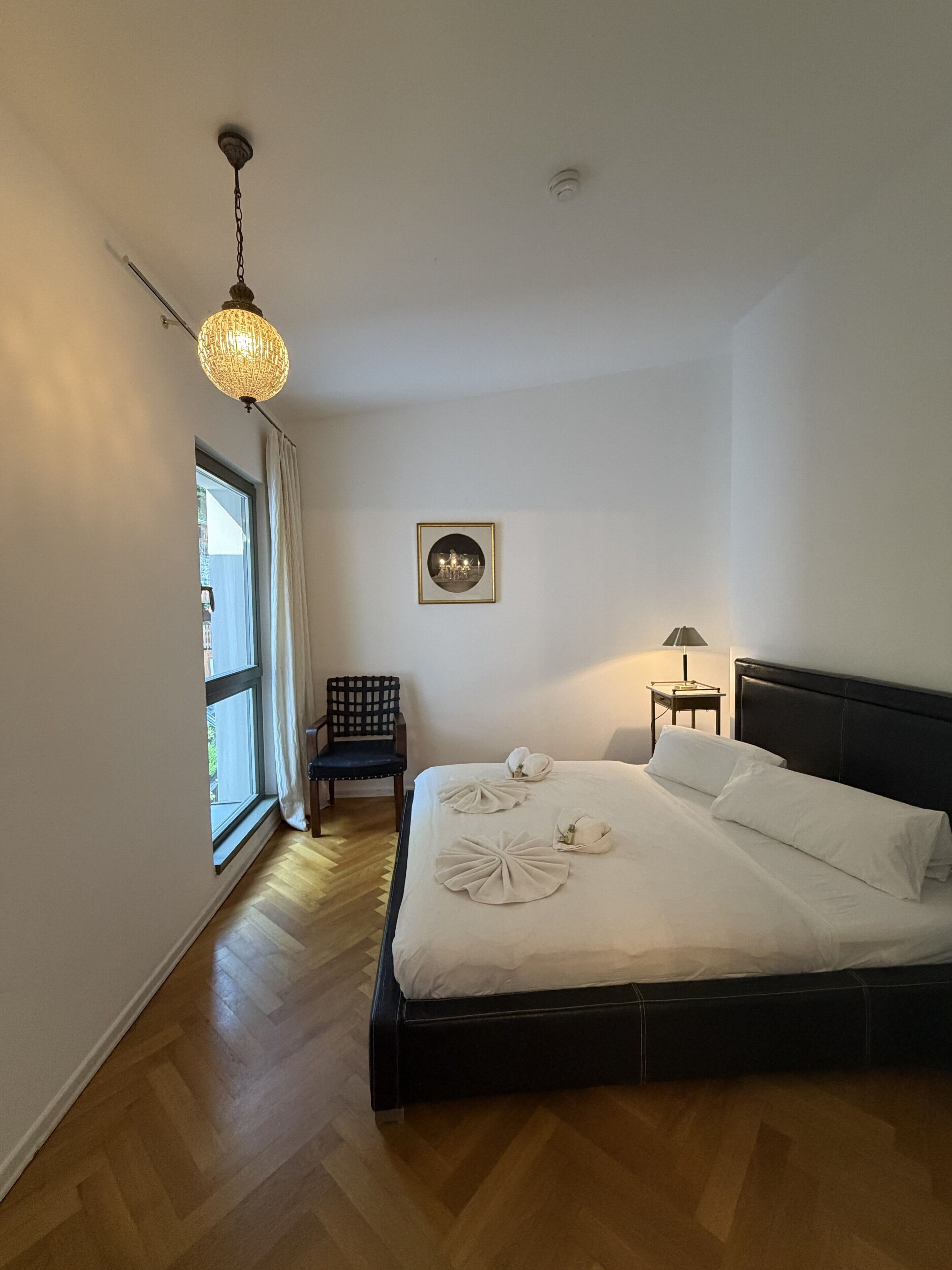 Une chambre à coucher avec un lit, une chaise, une lampe, un tableau au mur et une fenêtre.